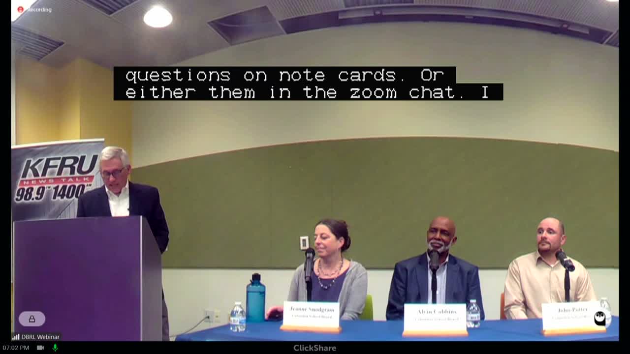A moderator stands next to three school board candidates sitting at a table.